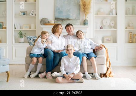 Portrait large family spending time together at home. They are happily sitting in the living room on the couch in a modern light interior. Caucasian parents with children spend time on weekends Stock Photo