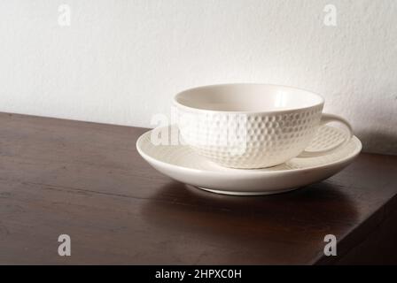 Beautiful empty white tea or coffee cup with rounded pattern texture as a golf ball, the cup is on the old wooden dark-brown table, white wall backgro Stock Photo