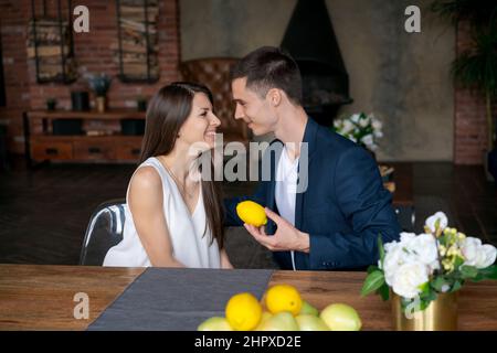 Romantic mature couple enjoying at home after office. Cheerful man and beautiful woman are hugging and giving each other a lemon fruit. Handsome man and woman spend evening together at home happy. Stock Photo