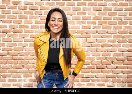 Trendy Hispanic female in leather jacket and with charming toothy smile looking at camera against brick wall in city Stock Photo