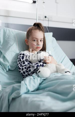 Unhealthy child in healthcare pediatric hospital patients treatment ward room while looking at camera. Sick kid holding teddybear and wearing oxygen tube while resting alone in pediatric clinic bed. Stock Photo