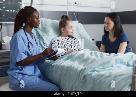 Healthcare pediatric clinic nurse taking notes about successful recovery while in patient room. Mother being attentive to nurse speaking about little girl health hazards while in recovery ward. Stock Photo