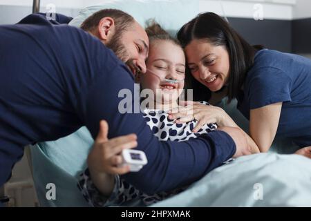 Happy young parents hugging hospitalized recovered little daughter while in pediatric clinic patient room. Cheerful family laughing and hugging in health pediatric hospital room. Stock Photo