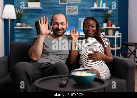 POV of interracial couple on videoconference call in living room while expecting together newborn at home. Cheerful people waving at relatives through modern video call communication. Stock Photo