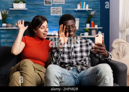 Smiling multiethnic couple waving at smartphone while in videoconference at home. Young married people discussing to their family through online videocall while sitting on sofa in living room. Stock Photo