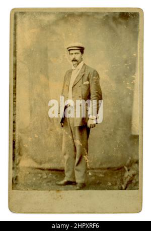 Original Victorian cabinet card portrait of a working class man wearing a flat cap, an ill-fitting suit, with too short sleeves, turned up collar, tie, taken outside with a canvas backdrop against a stone wall - late 1880's, early 1890's, U.K.