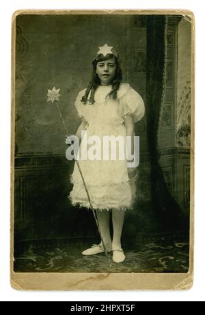 Original and charming Victorian cabinet card studio portrait of smiling older girl wearing a fairy costume with tinsel trim, holding a wand, she has large puff sleeves on a white party dress, fashionable circa 1895. Stock Photo