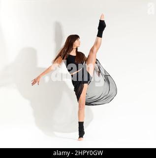 Full length of flexible female dancer with long brown hair in black clothes performing developpe pose against white background in studio Stock Photo