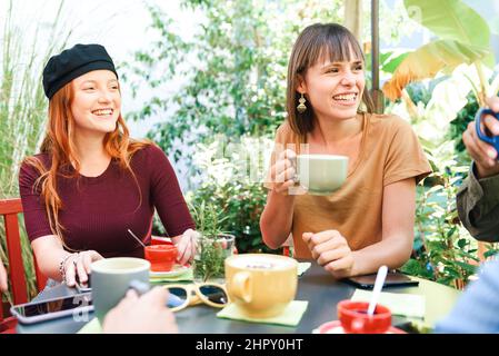 Group of happy friends with cups of hot coffee sitting at table in outdoor garden with green trees during break Stock Photo