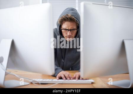 Burning the midnight oil. A young hacker working on dual screens and a laptop wearing a hoodie. Stock Photo