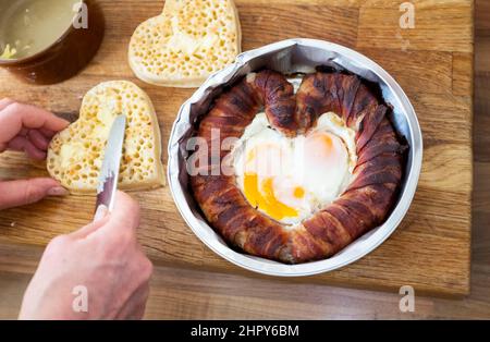 A quirky Valentines Day breakfast of heart shaped pigs in blankets or sausage and bacon with fried eggs with buttered crumpets