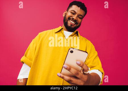 Surfing the net in the studio. Happy young man smiling cheerfully while using a smartphone to browse the internet. Carefree young man standing alone a Stock Photo