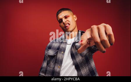 Young man winking and pointing at the camera in a studio. Handsome young man sticking his tongue out while standing against a red background. Fashiona Stock Photo