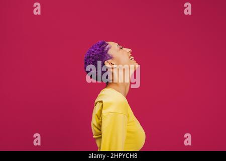 Purple haired young woman laughing cheerfully in a studio. Sideview of a fashionable young woman laughing with her eyes closed. Confident young woman Stock Photo