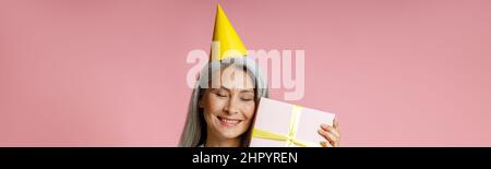 Happy mature Asian lady with yellow party hat holds gift box on pink background Stock Photo