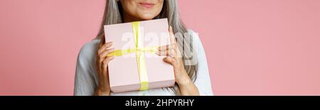 Positive mature female model holds gift box posing on pink background Stock Photo