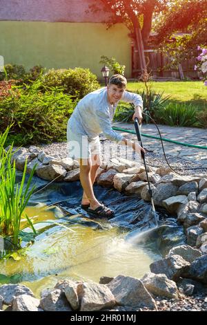 man cleans garden pond bottom with high-pressure washer from mud and sludge. Stock Photo