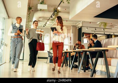 Three happy businesswomen walking through a modern office in the morning. Group of young businesswomen having a cheerful chat. Female entrepreneurs wo Stock Photo