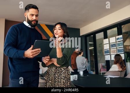 Two young businesspeople using a digital tablet while standing in a boardroom. Two business colleagues having a discussion during a meeting. Diverse b Stock Photo