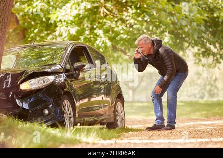 Mature Man Next To Car Crashed Into Tree Inspecting Accident Damage And Calling Emergency Services Stock Photo