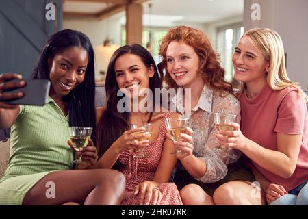 Multi-Cultural Group Of Female Friends Sitting On Sofa At Home Posing For Selfie Together Stock Photo
