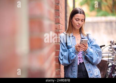 Young Woman Standing By Wall Looking At Mobile Phone Checking Text Messages And Social Media Stock Photo