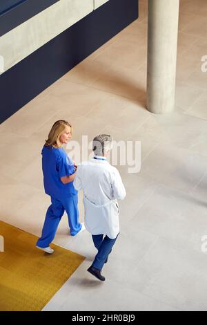 Overhead Shot Of Male And Female Doctors In White Coats And Scrubs Walking Through Busy Hospital Stock Photo