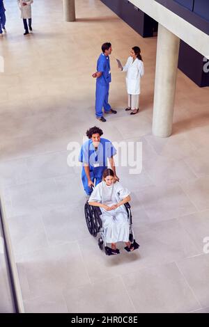 Overhead Shot Of Nurse Wearing Scrubs Pushing Female Patient In Wheelchair Through Hospital Building Stock Photo