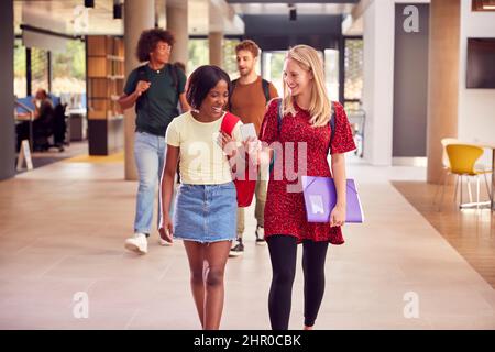 Two Female Students In Busy University Or College Building Looking At Mobile Phone Stock Photo