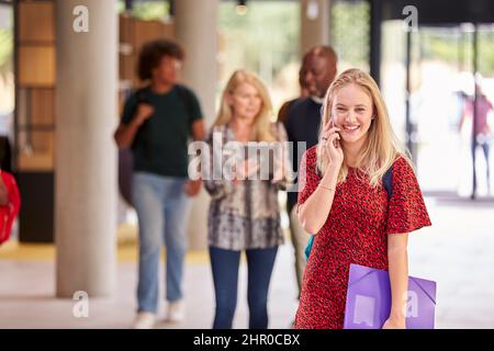 Female Student Talks On Mobile Phone As She Walks Through Busy University Or College Building Stock Photo