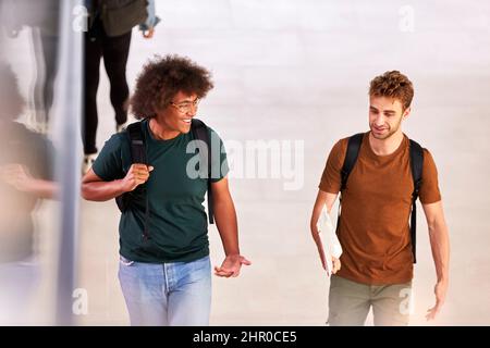 Two Male Students In Busy University Or College Building Talking As They Walk Along Corridor Stock Photo