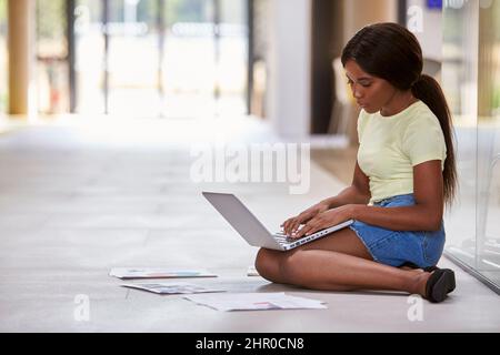 Female University Student Sitting On Floor Of College Building Using Laptop Stock Photo