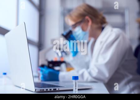 Female Lab Worker Wearing Lab Coat Working On Covid-19 Vaccine With Laptop And Microscope Stock Photo