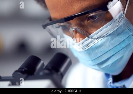 Close Up Of Female Lab Worker Wearing PPE And Safety Glasses Looking Through Microscope Stock Photo