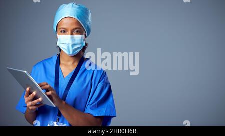 Studio Portrait Of Female Surgeon Wearing Scrubs And Face Mask Using Digital Tablet Stock Photo