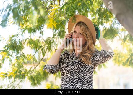 Gardener woman with a straw hat talks on the phone under a mimosa in the garden Stock Photo