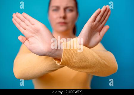 Frightened angry caucasian woman in yellow sweater crossing hands makes stop gesture, demonstrates rejection isolated over blue background. Stock Photo