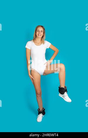 Fit woman smiling wearing white bodysuit, sneakers and black wrist weights doing exercises to be in good shape on blue background. Healthy lifestyle Stock Photo