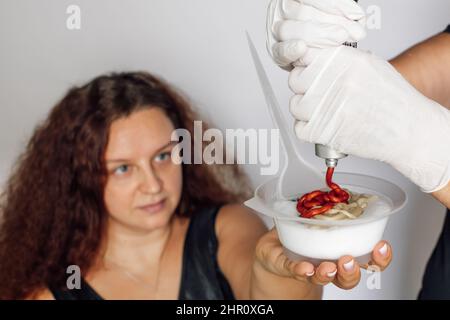 Long lush dark red haired woman with transparent bowl full of hair dye in hand looking at latex gloved hands squeezing content of dyeing tube into Stock Photo