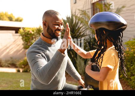 Happy african american girl wearing helmet giving high-five to father in backyard on sunny day Stock Photo