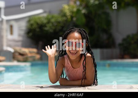 Portrait of happy african american girl wearing sunglasses leaning at poolside waving on sunny day Stock Photo