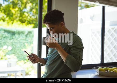 Young biracial man drinking coffee from cup while using smartphone in kitchen at home Stock Photo