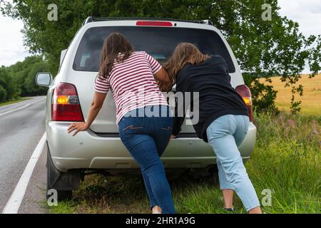 A broken car. Two young women push a broken car on the road, a breakdown, against the background of a yellow field. Stock Photo