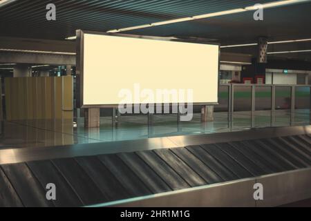 A blank advertising poster in an international airport near an empty moving luggage belt. Baggage claim area with a long horizontal advertisement bann Stock Photo