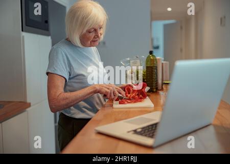 Pensioner female using laptop while cooking at home Stock Photo