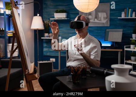 Senior sketch artist using virtual reality glasses doing air gesture improving freehand technique in creative workshop. Retired art teacher with modern technology vr goggles looking at 3d artwork. Stock Photo