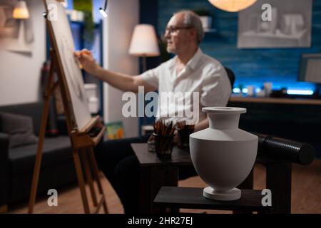 White model vase on wooden table used for light study by retired man artist drawing using sharp pencil on easel in home art studio. Selective focus on still life object used as subject for sketch. Stock Photo