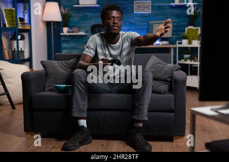 Upset sad player making furious gesture after losing virtual gaming competition sitting on couch in living room. Pro gamer playing online video games using wireless joystick. Cyber space Stock Photo