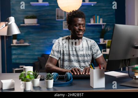 Smiling freelancer man browsing information on internet working at business project. African american businessman sitting at desk table analyzing financial graphs on computer. Remote work Stock Photo
