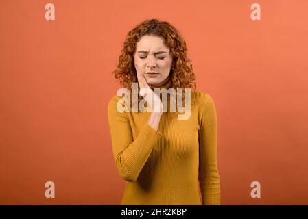 Young and attractive caucasian redhead girl touching her mouth with toothache isolated on orange studio background. Stock Photo